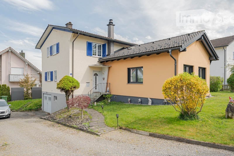 Two-story house with a cream-colored facade, blue shutters, and a gray garage. The house features a well-maintained lawn, small trees, and a paved driveway.