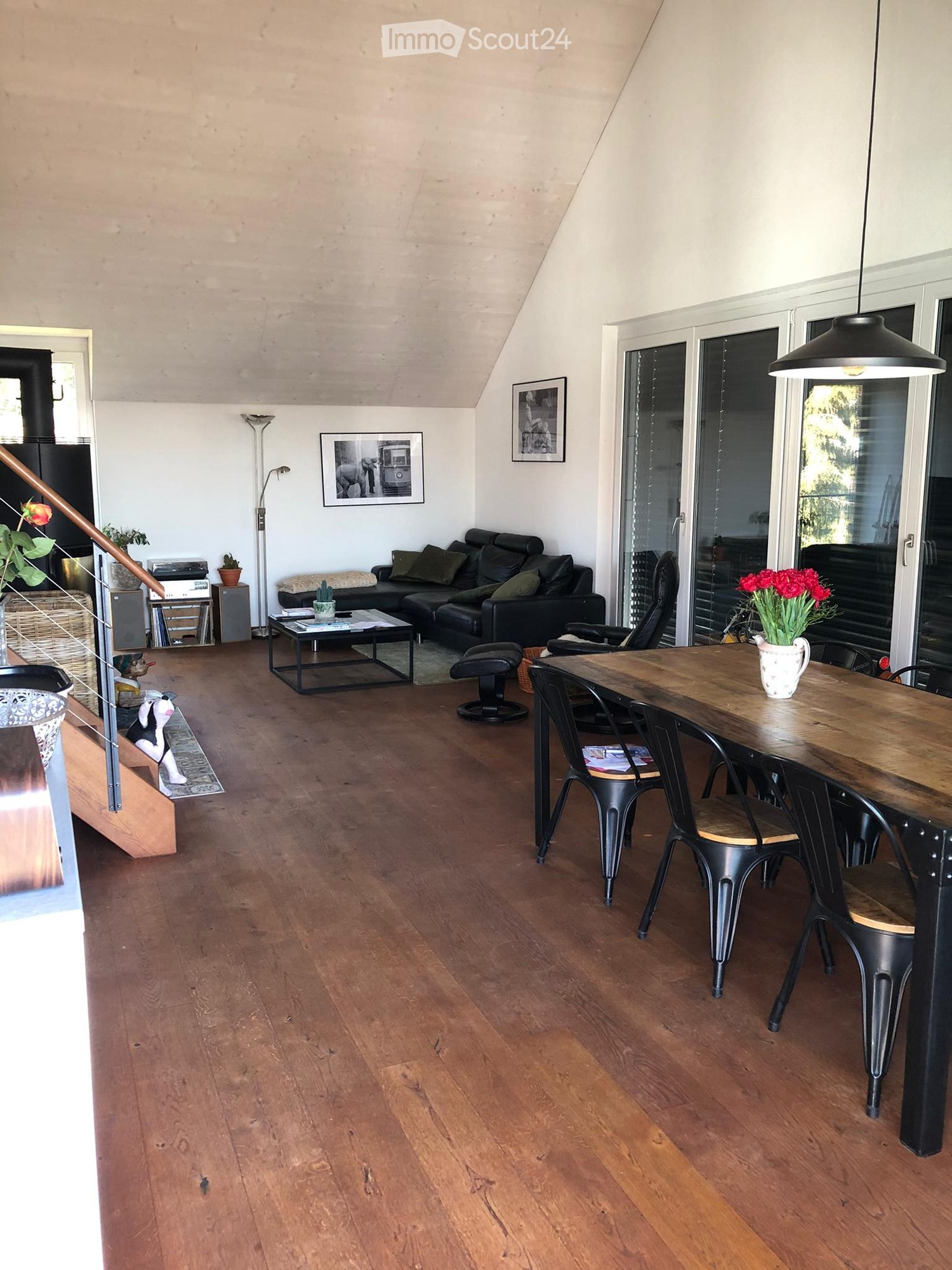 Open-plan living and dining area with wooden flooring, a black leather sofa, a coffee table, a dining table with chairs, and decorative items including a flower vase and picture frames.