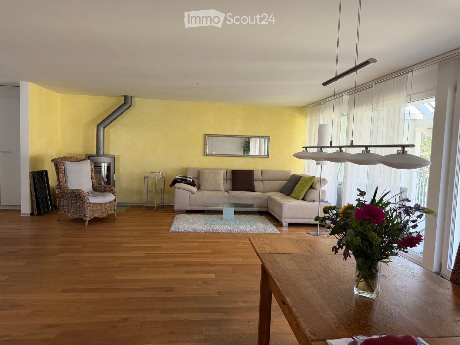 Bright living room with wooden flooring, a beige sectional sofa, a wicker armchair, a glass coffee table, a large flower vase on a wooden side table, and a modern floor lamp.