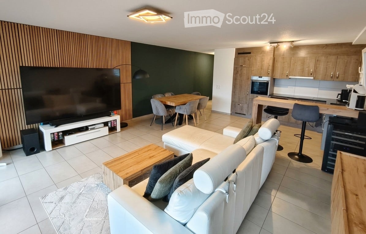 Modern open-plan living and dining area with a white leather sofa, wooden coffee table, flat-screen TV on a stand, dining table with chairs, and a kitchen area with cabinets and appliances.