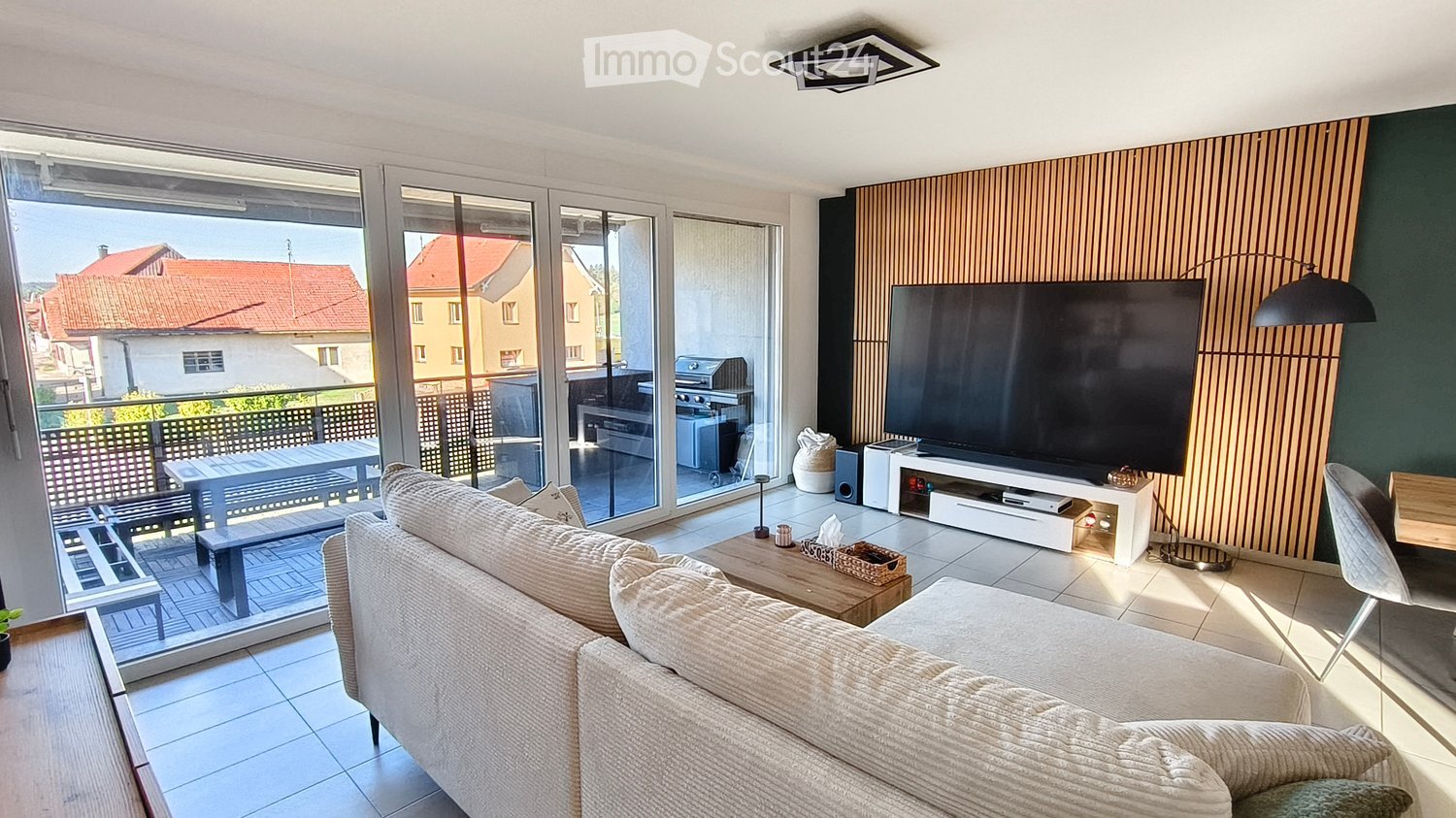Modern living room with large sliding glass doors leading to a balcony, a cream-colored sofa, a wooden coffee table, a flat-screen TV on a white stand, and a dining area with a table and benches.