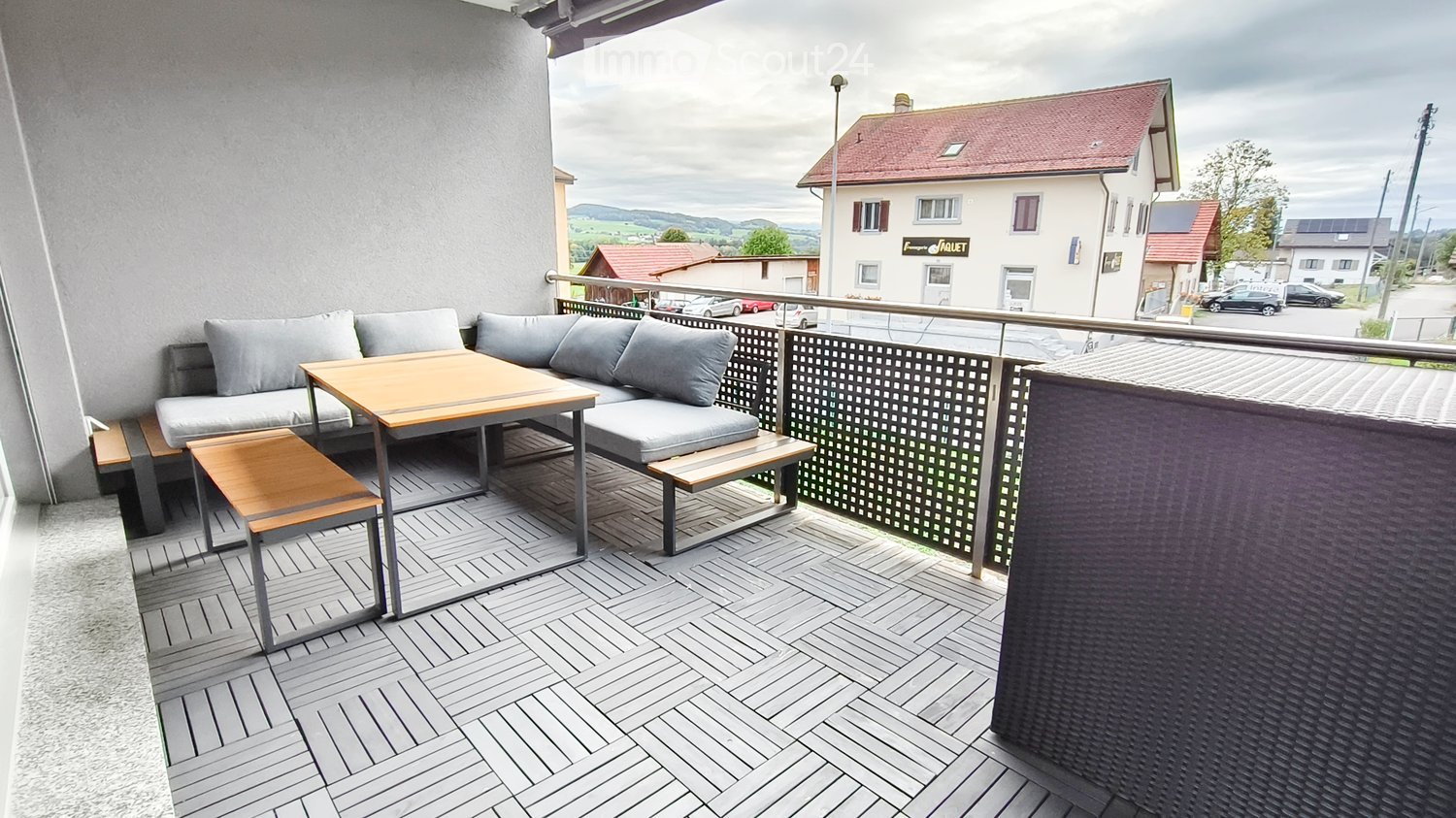 Balcony with grey sofa, wooden table, and two stools, overlooking a town with houses and a mountainous backdrop.