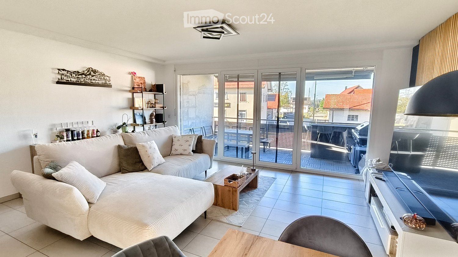Modern living room with a large sectional sofa, coffee table, TV, and shelves. Large glass sliding doors open to a balcony with outdoor furniture.