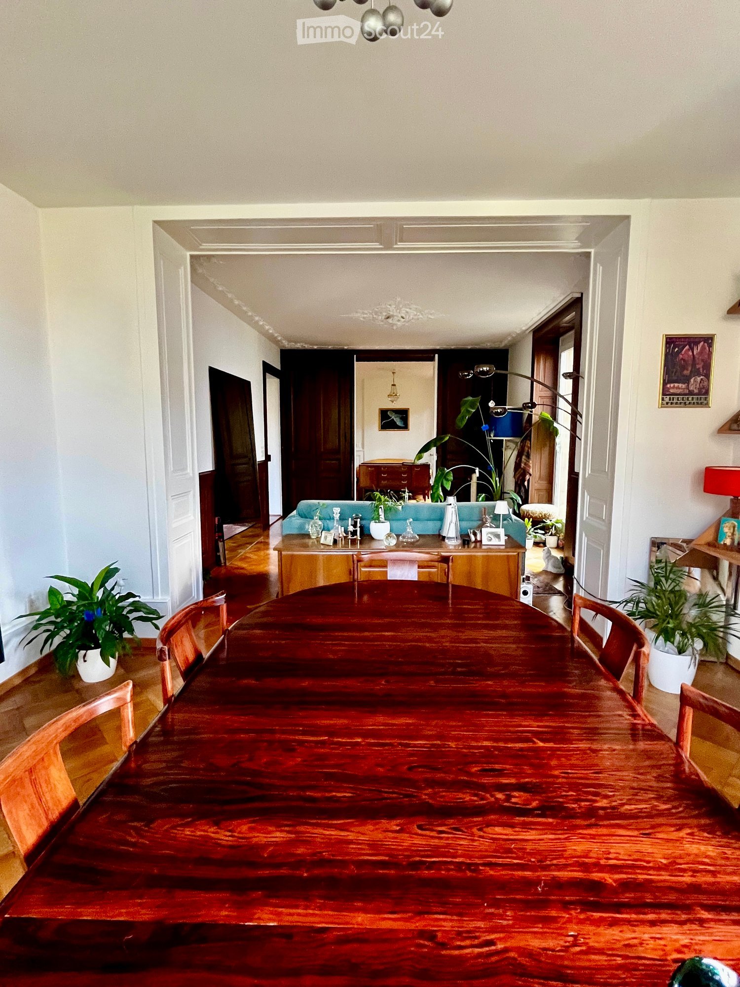 A well-lit dining area with a large wooden dining table and chairs, a blue sofa in the background, and decorative items on a side table.