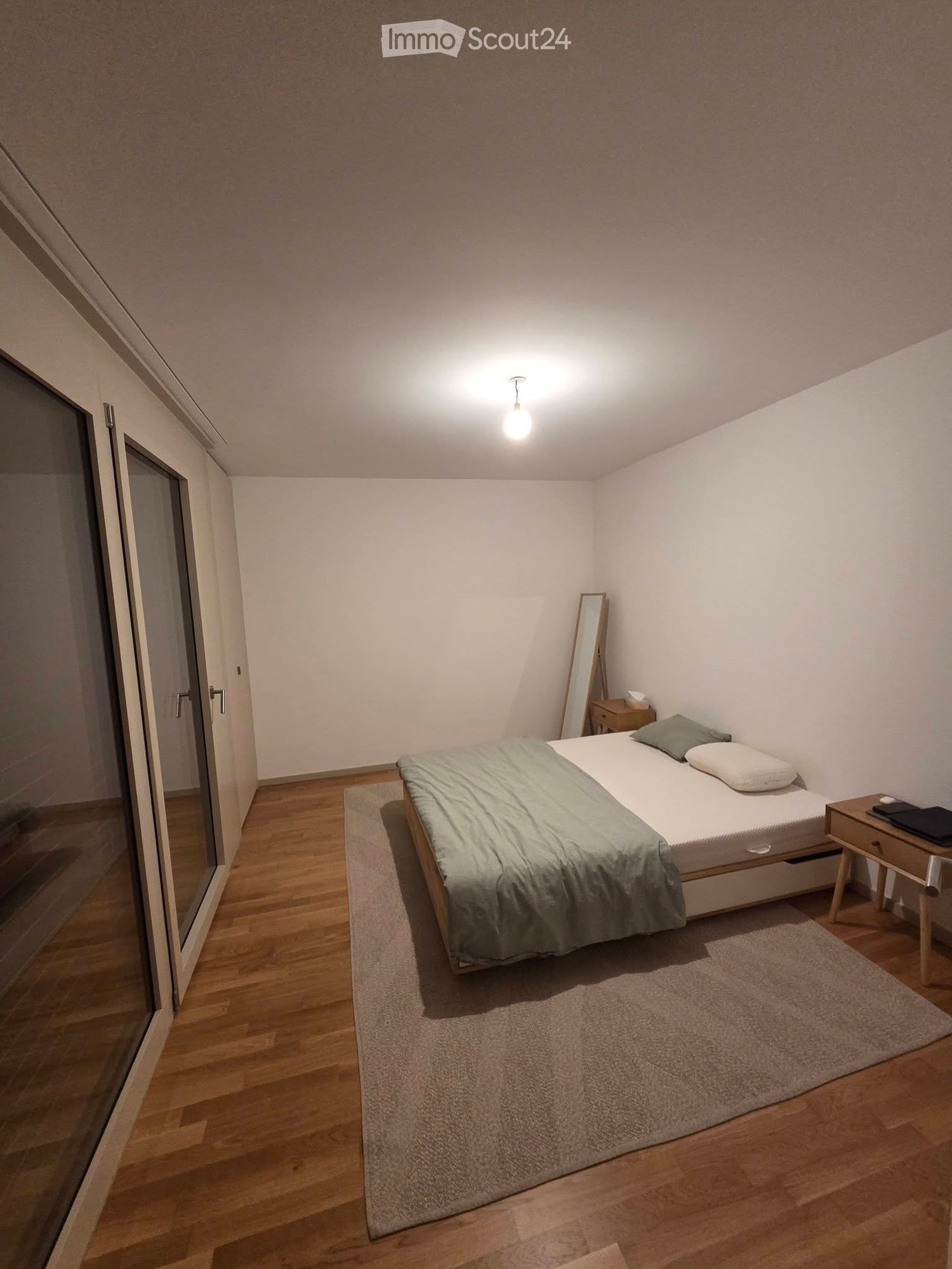 Modern bedroom with a single bed, nightstand, mirror, and laptop on the nightstand. The room features wooden flooring and a large window with a glass door.