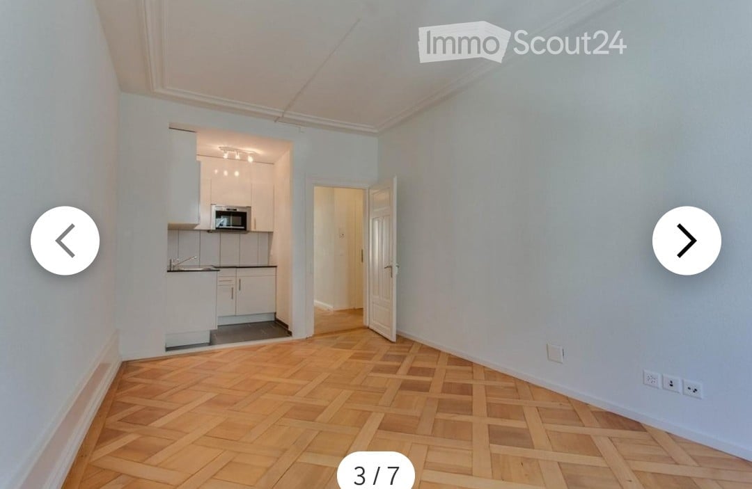 Empty room with light blue walls and wooden herringbone flooring. An open doorway leads to a small kitchen area with white cabinets and a microwave.