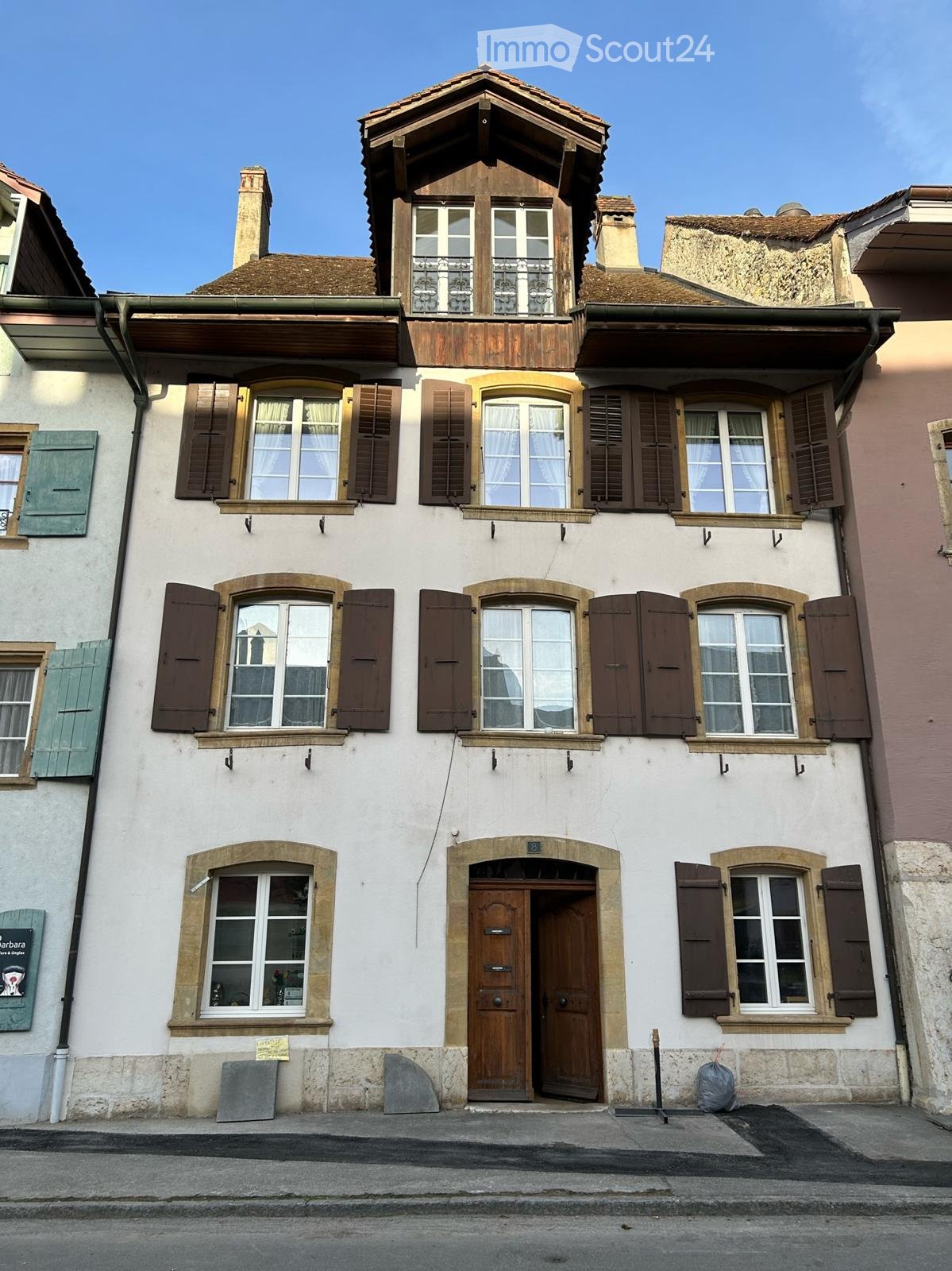 A three-story residential building with white walls, multiple windows with shutters, and a wooden front door. The roof features a small attic with dormer windows and a balcony.
