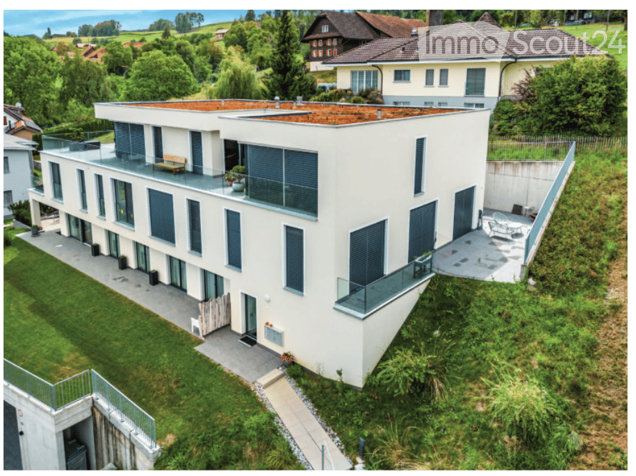 Modern two-story house with white exterior, large windows, balconies, and a flat roof. The house is surrounded by a well-maintained garden with greenery and a pathway leading to the entrance.