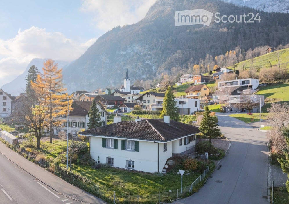 A picturesque village scene with traditional houses, a church tower, and surrounding mountains under a clear blue sky.