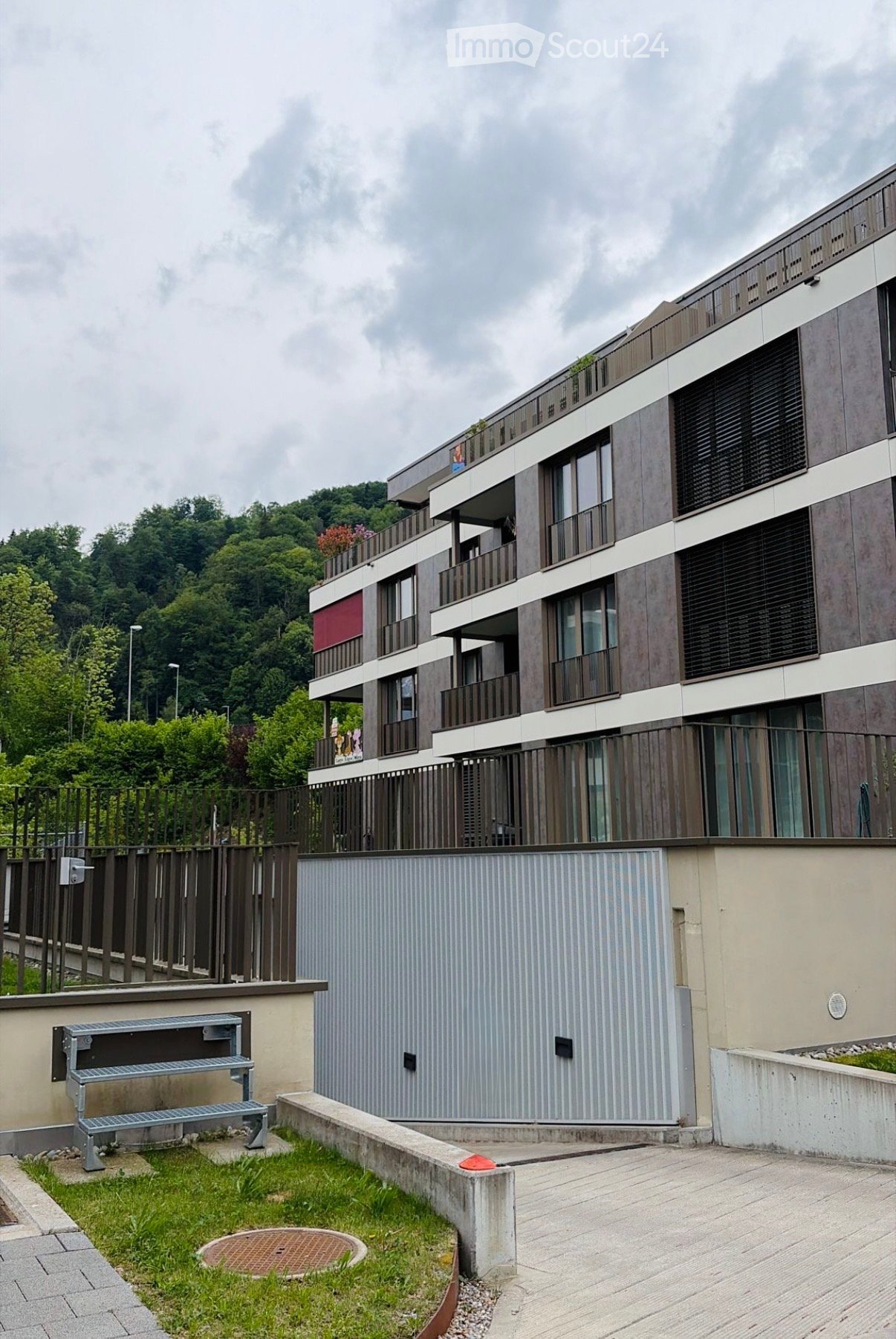 Modern multi-story residential building with balconies, large windows, and contemporary design, surrounded by greenery and a small garden area with a metal staircase.