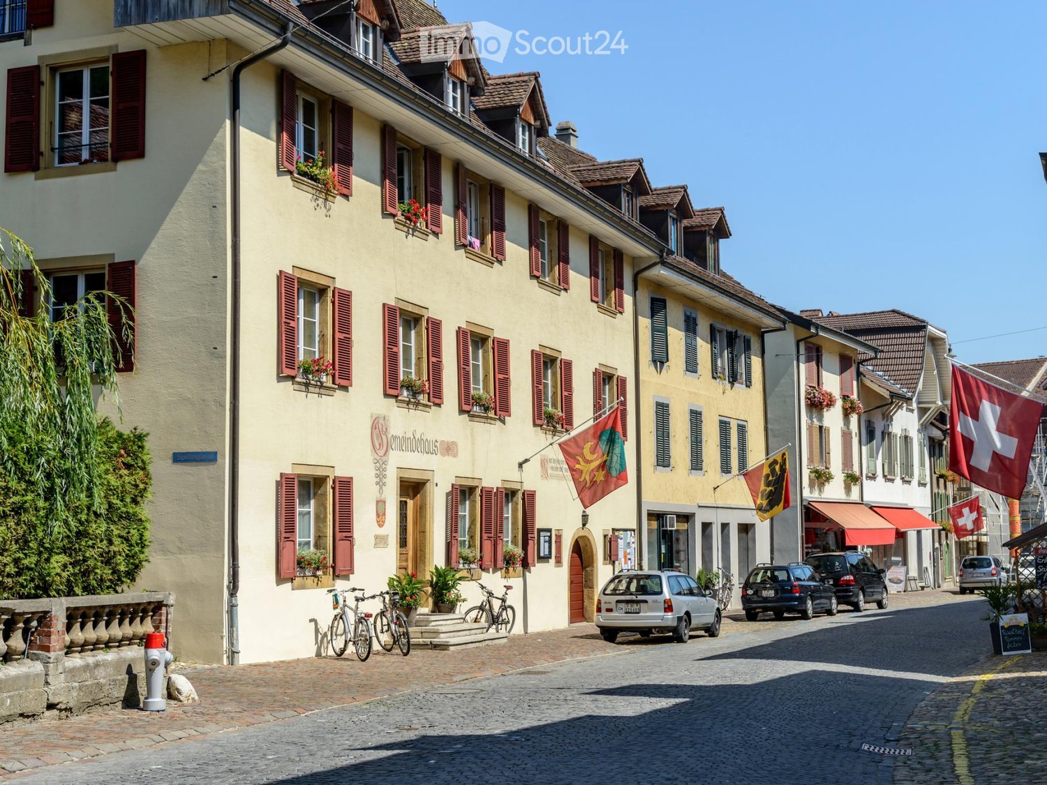 A charming European street lined with multi-story buildings featuring red shutters, flower boxes, and Swiss flags. The street is paved with cobblestones and has parked cars and bicycles.