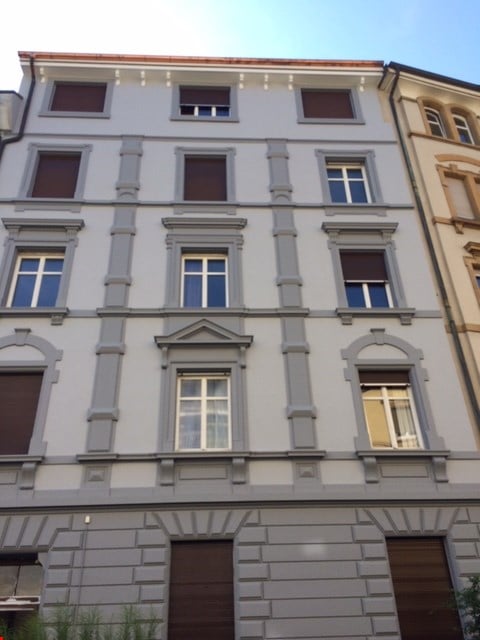 Four-story residential building with grey facade, multiple windows, and decorative pilasters.