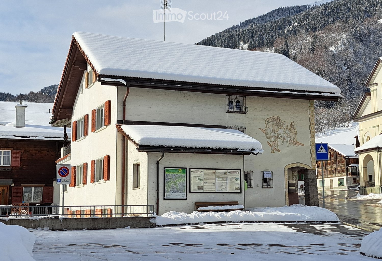 A two-story building with snow-covered roof and red shutters, located in a snowy village with mountains in the background.