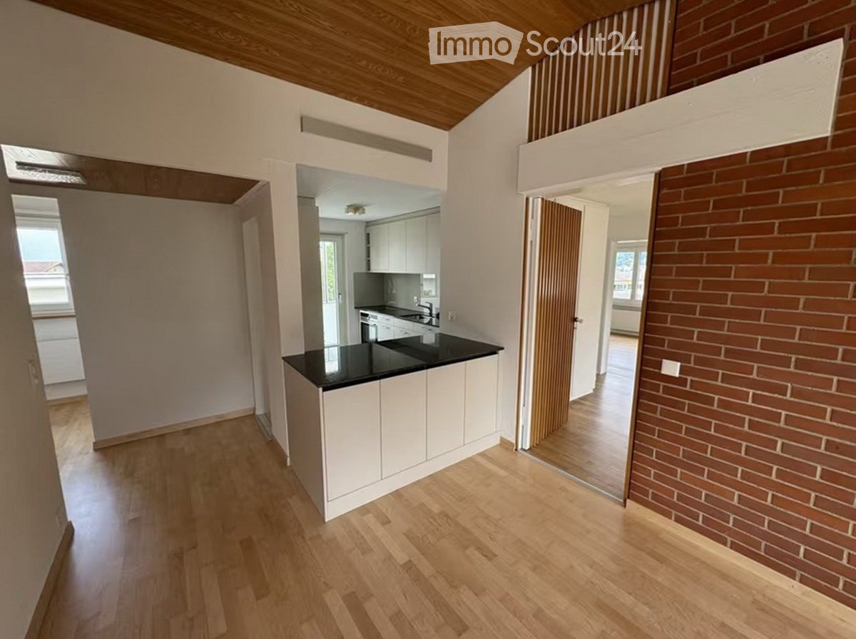 Open-plan kitchen area with white cabinets, black countertop, and a central island. Wooden flooring and exposed wooden ceiling beams.