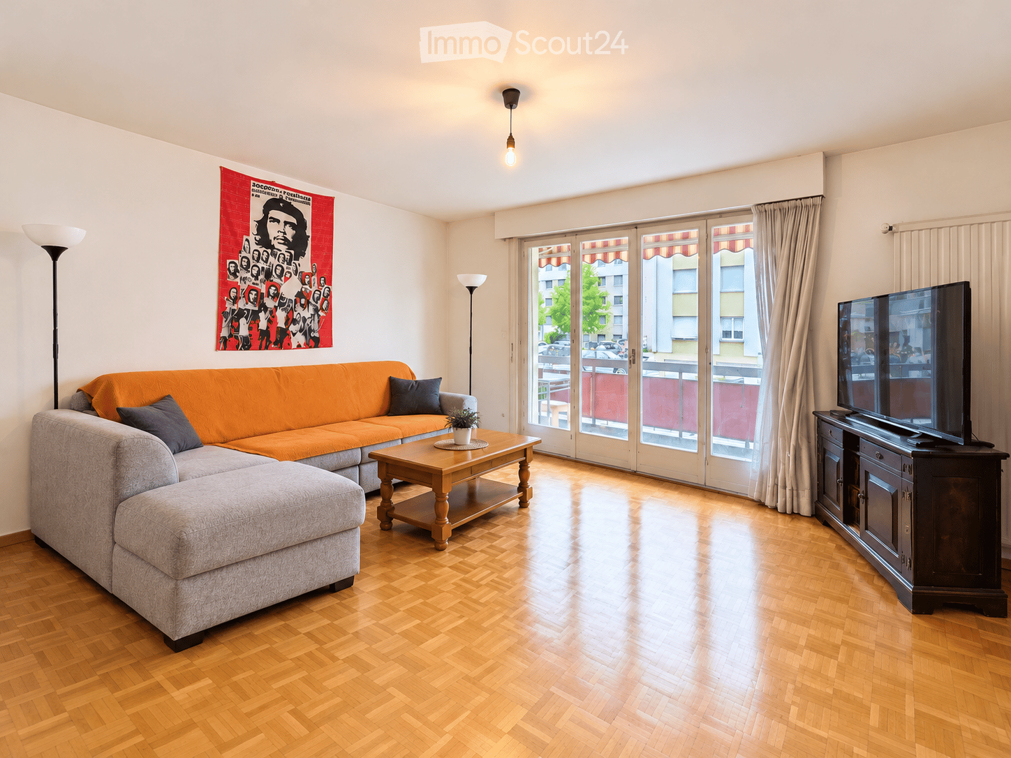 Living room with a grey sectional sofa, orange throw blanket, coffee table, TV, floor lamps, and a potted plant on the coffee table. Large windows with curtains lead to a balcony.
