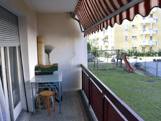Balcony with a small table, stool, broom, and a view of a playground and residential buildings.