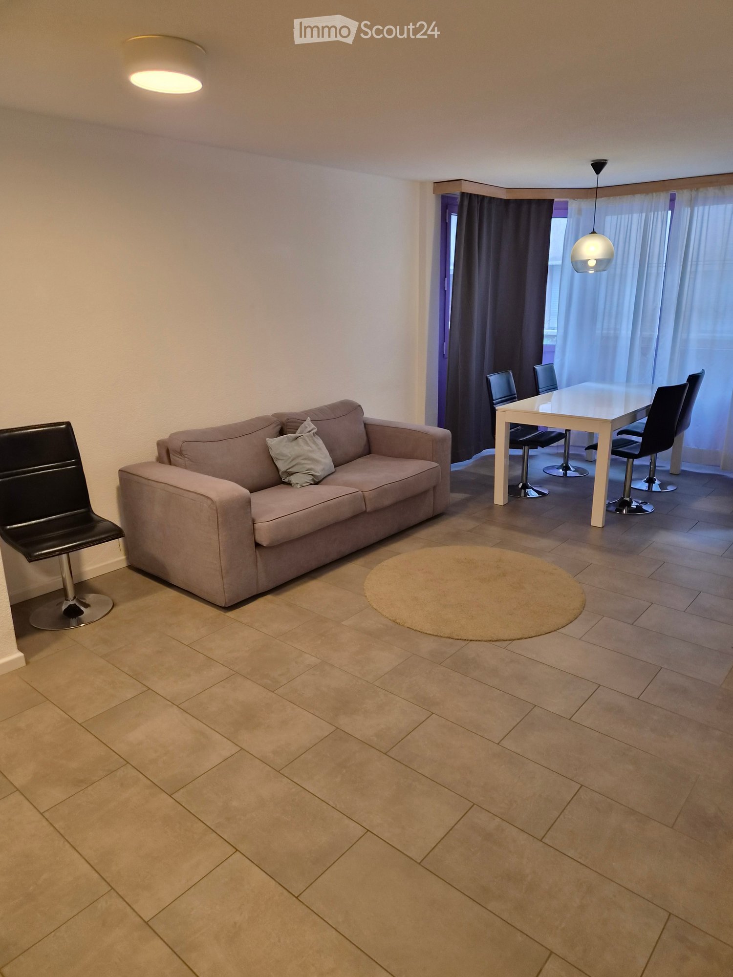 Living and dining area with a beige sofa, a round rug, a white dining table with four chairs, and a black bar stool near the sofa.