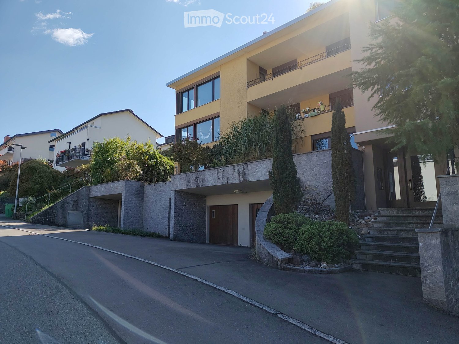 Modern two-story house with yellow facade, large windows, and a garage. The house is surrounded by greenery and has a concrete retaining wall with steps leading up to the entrance.