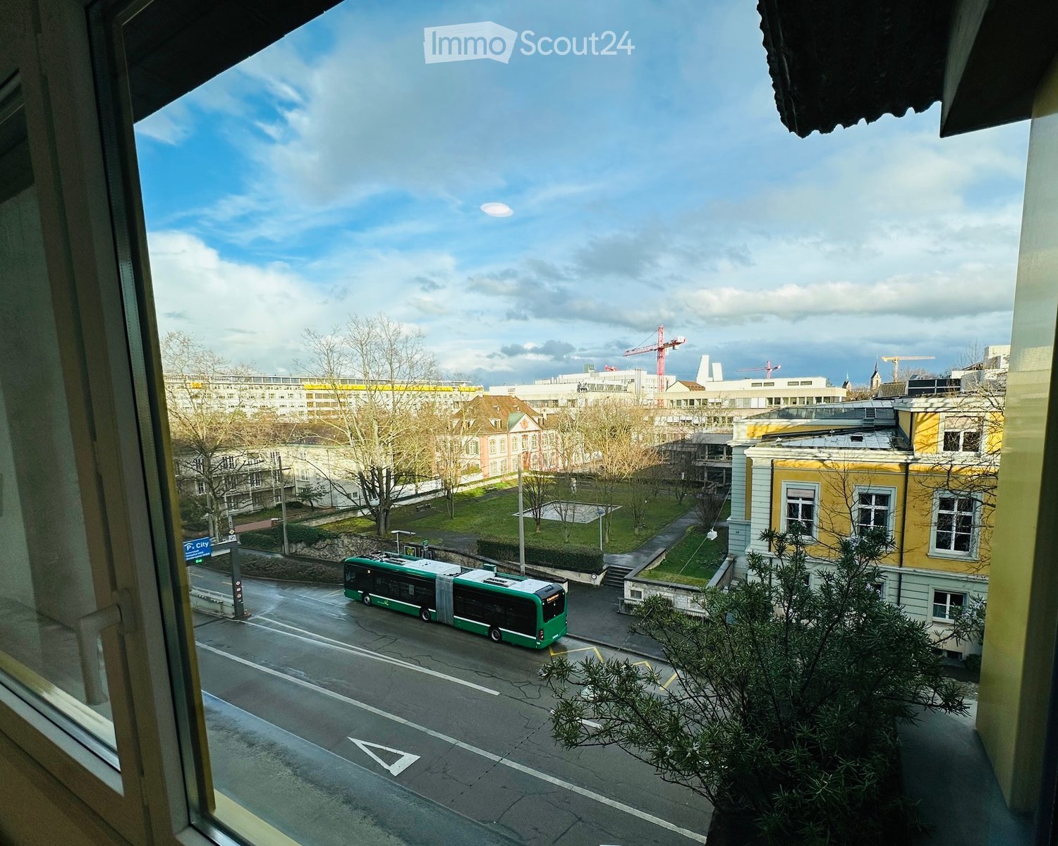 Balcony view overlooking a city street with a green and white bus, buildings, trees, and a clear sky.