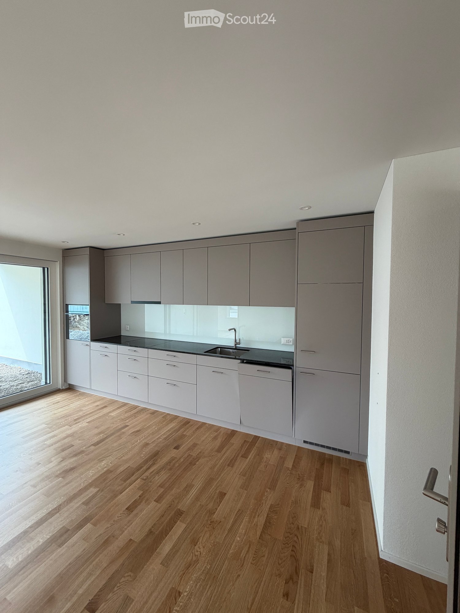 Modern kitchen with light grey cabinets, white countertops, and wooden flooring. Features a large window and built-in sink with a modern faucet.