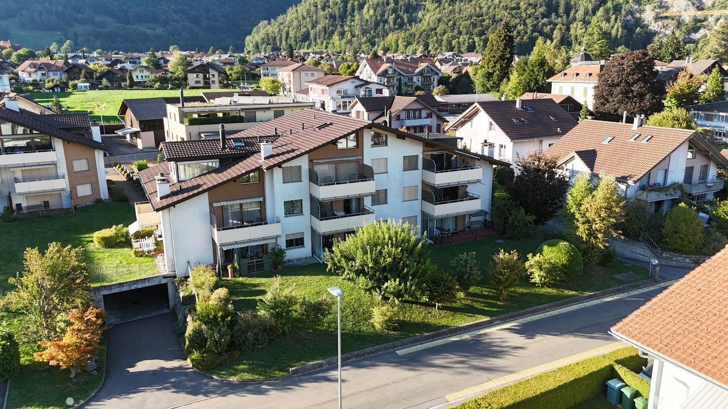 Aerial view of a multi-story residential building with a brown tiled roof, surrounded by greenery and neighboring houses in a suburban area.