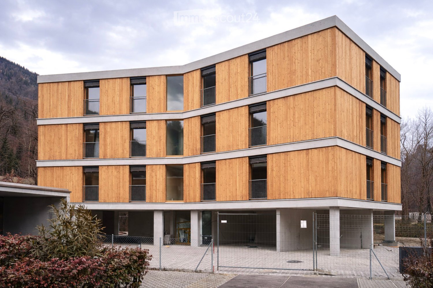 3 story wooden-paneled building, windows with balcony, fenced off entrance, ground floor with concrete pillars