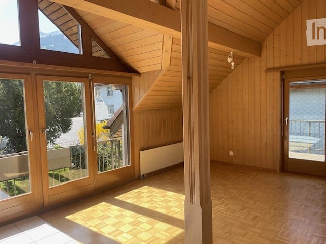 Bright, empty room with wooden floors and walls, large windows and sliding doors leading to a balcony, visible mountain view outside.