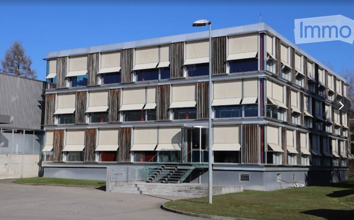 Modern multi-story building with a clean, geometric design, featuring large windows with roller shades and a raised entrance with steps.