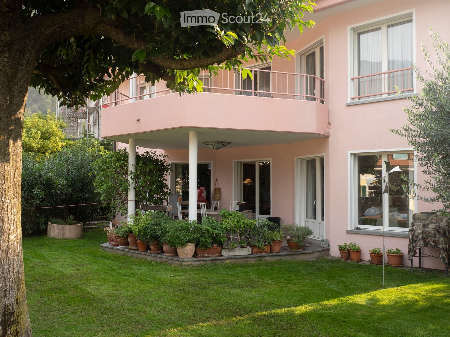 A well-maintained garden with a lawn, potted plants, and a raised patio area with outdoor furniture. The house features pink walls, large windows, and balconies.