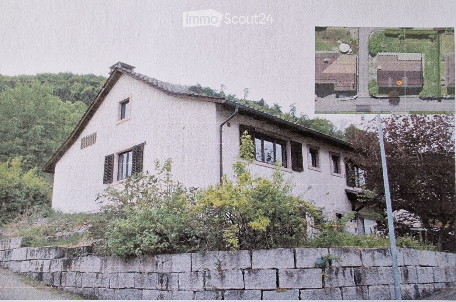 A two-story house with a sloped roof, multiple windows, and a stone retaining wall in front. The house is surrounded by greenery and trees.