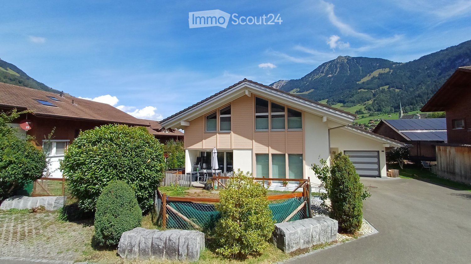 A two-story house with a modern facade, surrounded by greenery and mountains in the background. Features include a garage, patio area, and outdoor seating.