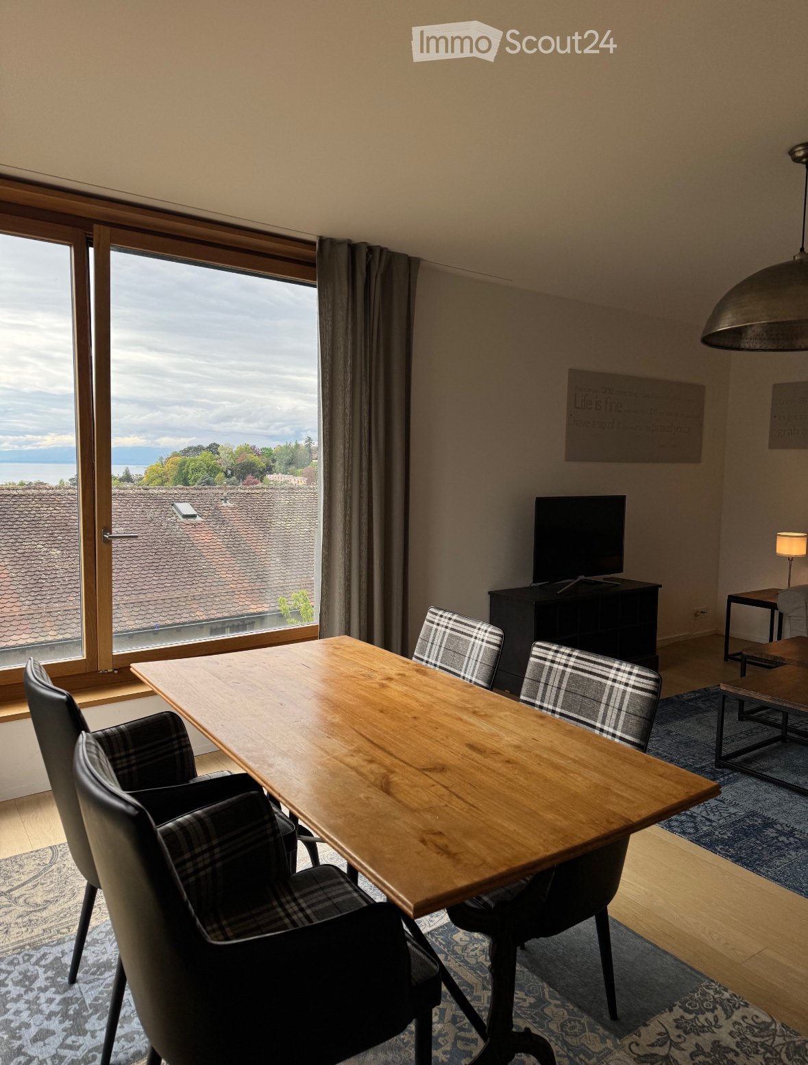Bright dining area with a wooden table, four chairs, a TV on a stand, and a lamp, with large windows offering a scenic view.