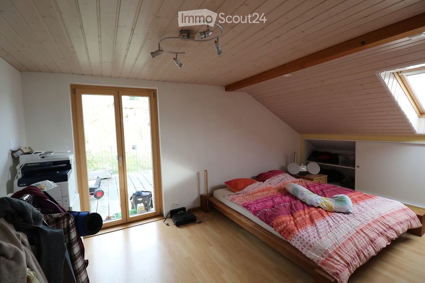 Bedroom with wooden floor, slanted ceiling, skylight, large window, bed with colorful bedding, nightstand, printer, chair, and built-in cabinet.