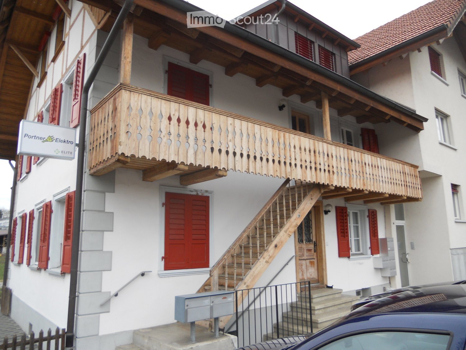 Two-story residential building with white walls, red shutters, and a wooden balcony on the upper floor. A wooden staircase leads to the entrance, and a car is parked in front.