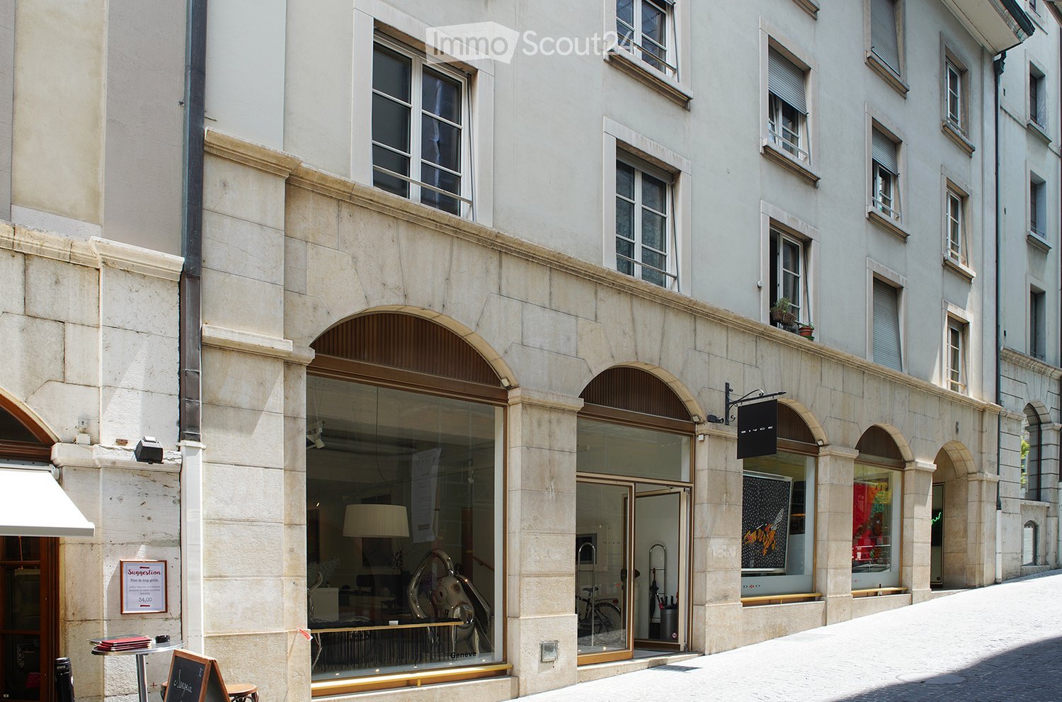 Classic European building facade with arched windows and doors, featuring a storefront with glass display windows and a small outdoor seating area.