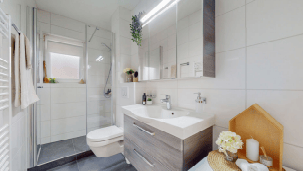 Modern bathroom with white tiles, a large mirror, a sink with a modern faucet, a shower, a toilet, and a wooden cabinet. A towel hangs on the left side, and a small plant is placed on the windowsill.