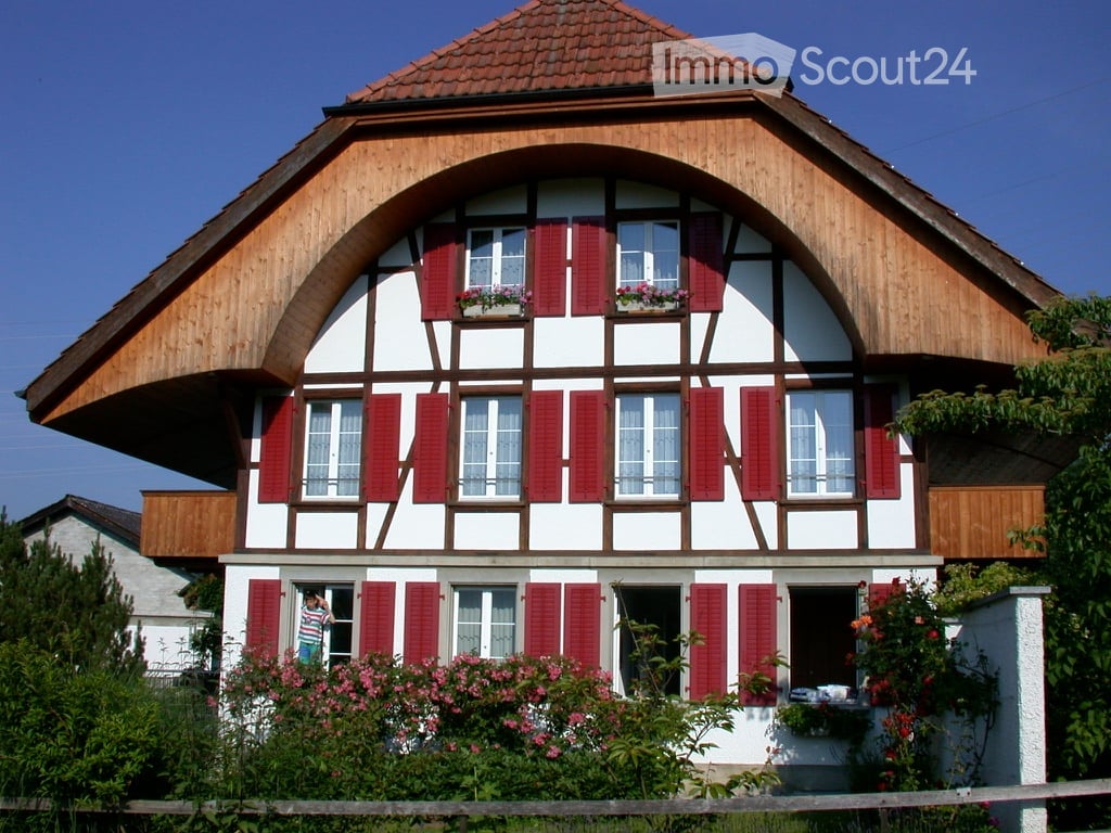 Traditional two-story house with white walls, red shutters, wooden beams, and flower boxes on the windows.