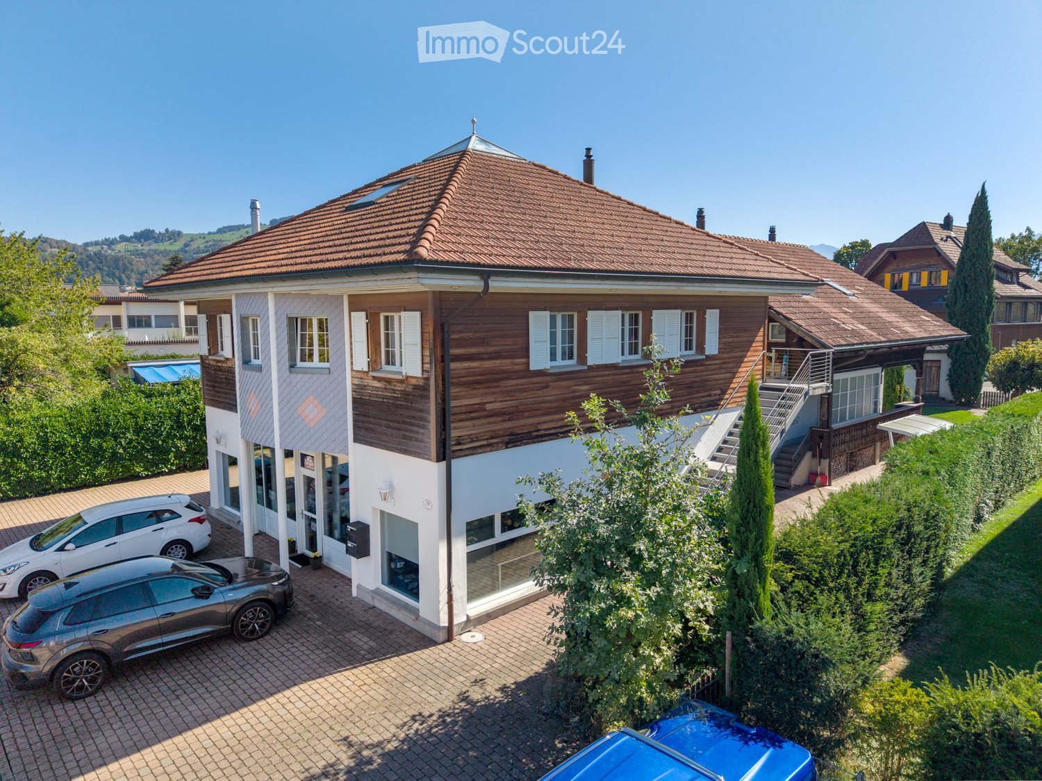 A two-story house with a red-tiled roof, white and wooden exterior, multiple windows, and a small staircase leading to an upper level. The house is surrounded by greenery and has a driveway with parked cars.