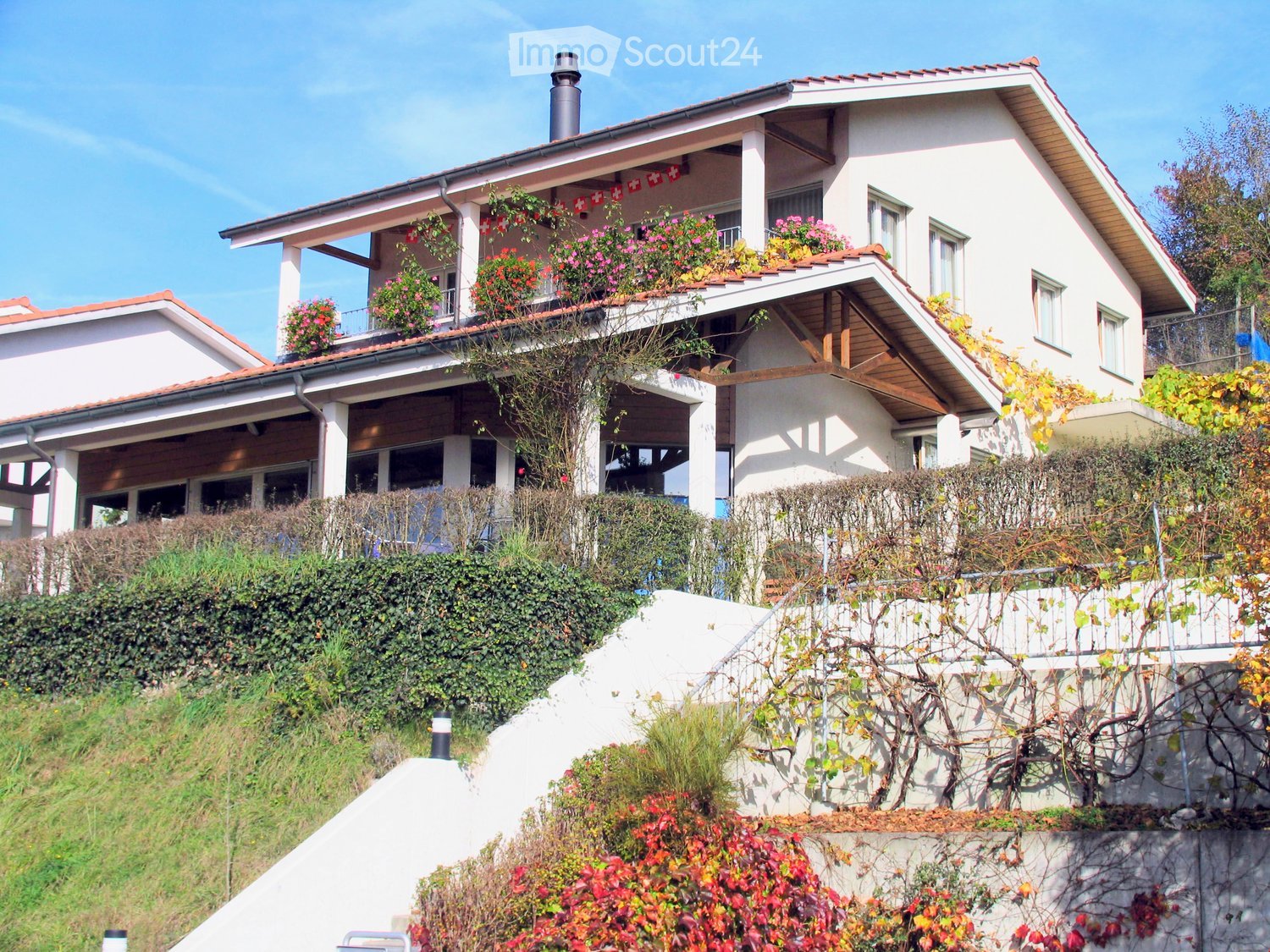 Two-story house with white walls, red-tiled roof, balcony with flower boxes, and surrounding greenery.