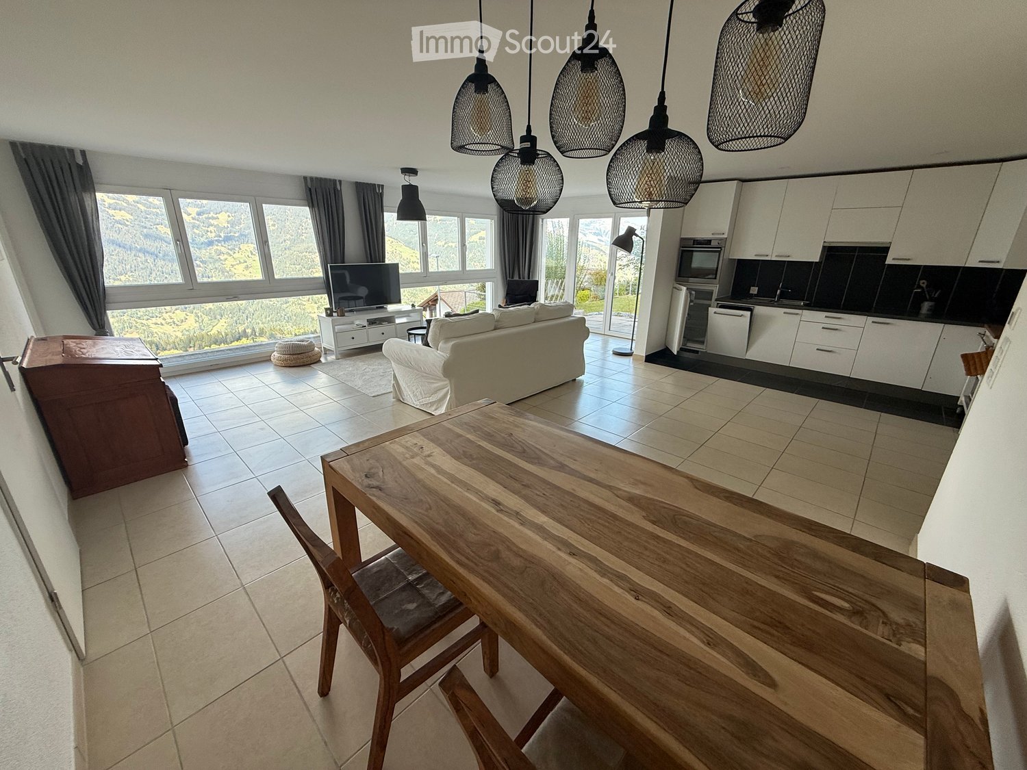 Open-plan living and dining area with a view, featuring a sofa, dining table, chairs, TV, and a modern kitchen with cabinets and appliances.
