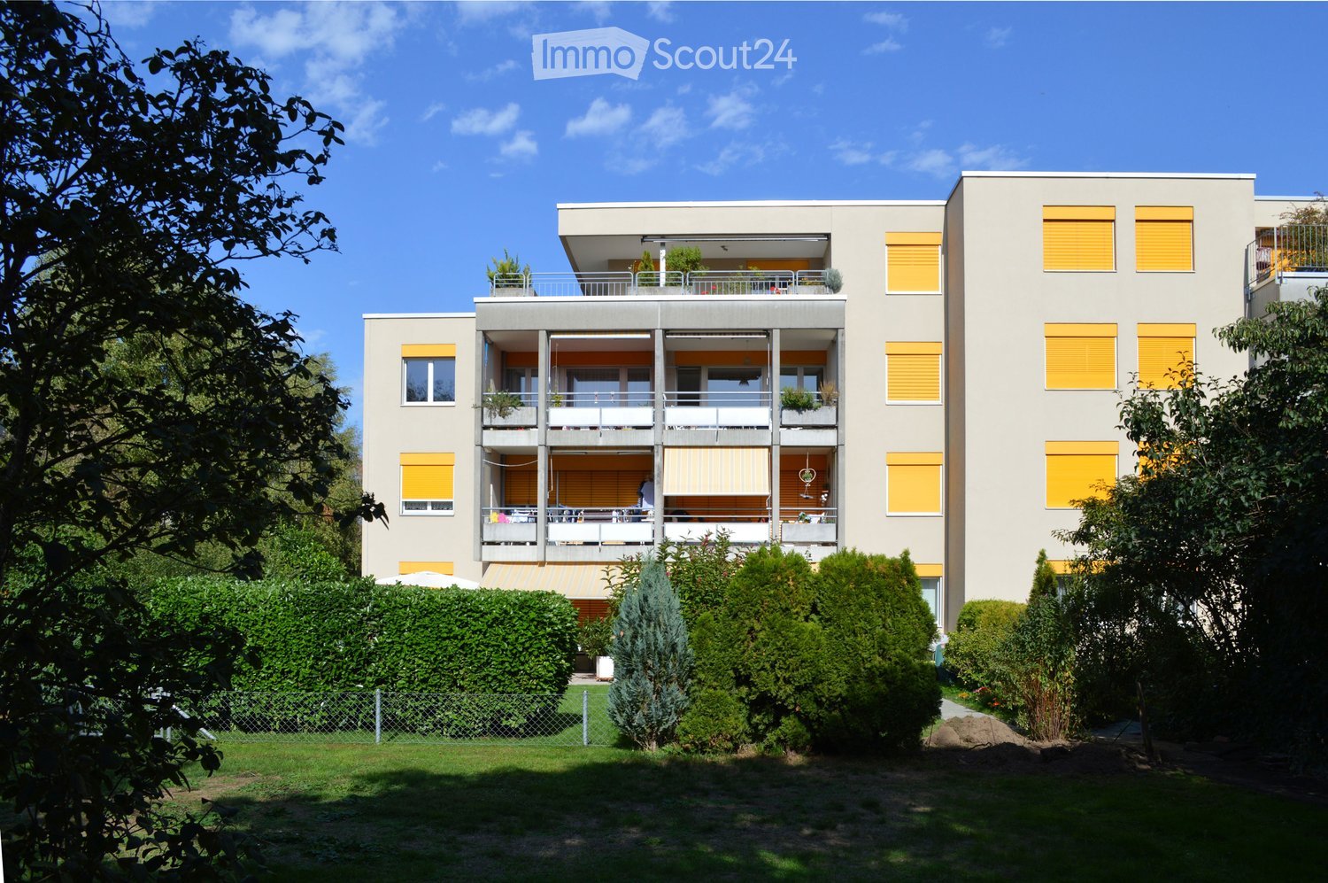 Modern multi-story residential building with balconies, yellow shutters, and surrounding greenery.
