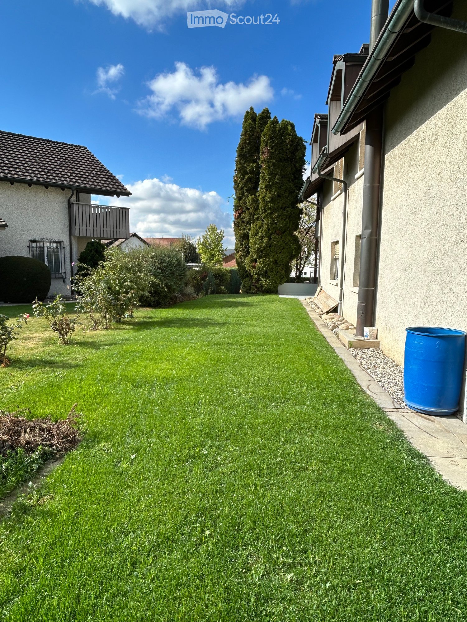 Well-maintained lawn with trimmed grass, small garden beds, and a blue water barrel near the house.