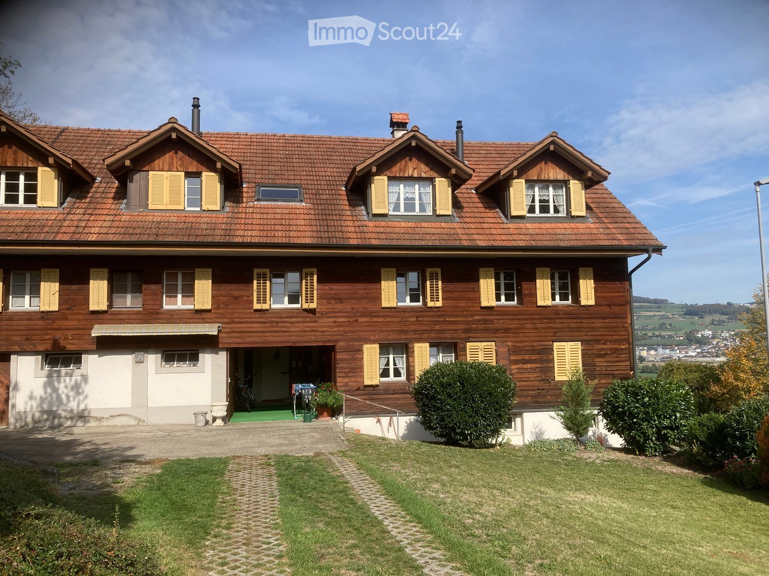 Two-story wooden house with red-tiled roof, multiple windows with yellow shutters, and a small garage. The house is surrounded by a well-maintained lawn and garden with bushes and a paved pathway leading to the entrance.