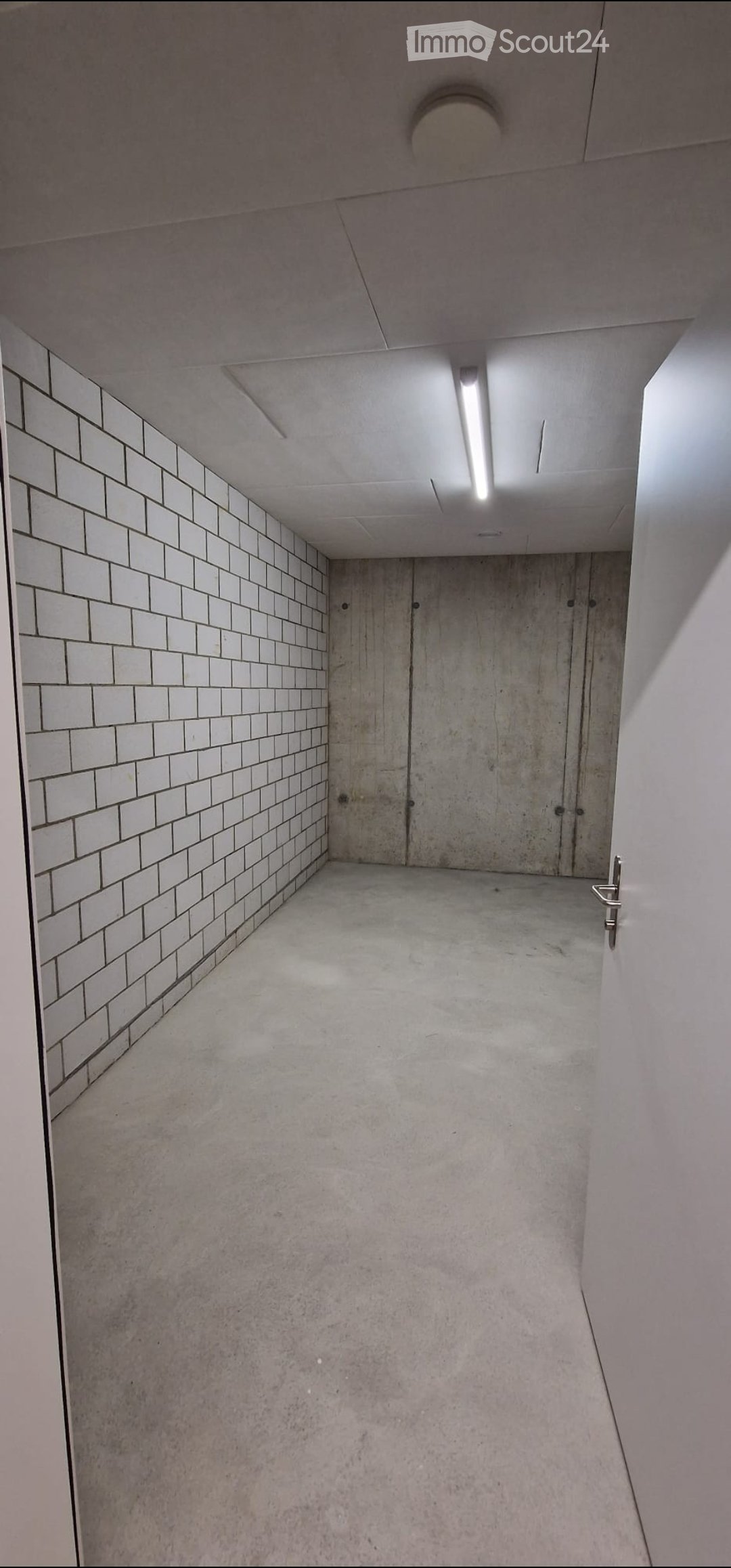 Modern, minimalist hallway with white tiled walls, concrete floor, and recessed lighting. Features a closed door at the end and a metallic handle on the right side.