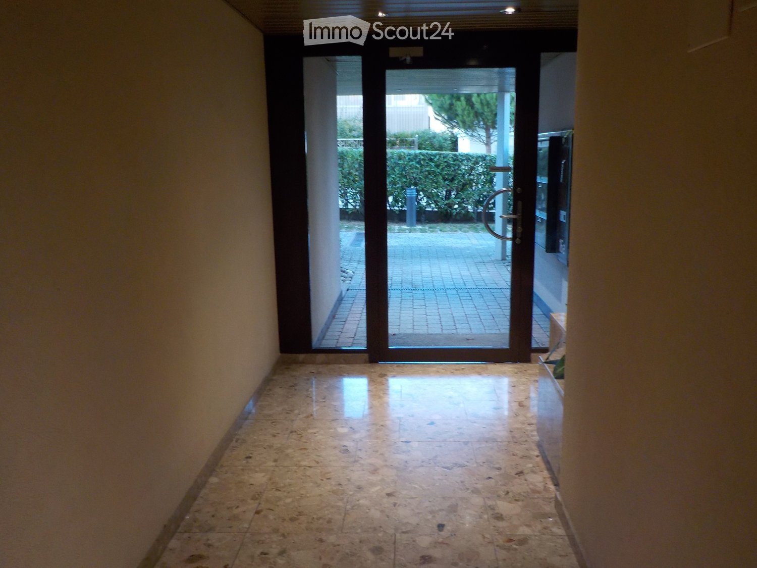 A clean, well-lit hallway with a polished tile floor, leading to a glass door that opens to an outdoor area with a paved patio and greenery visible beyond.