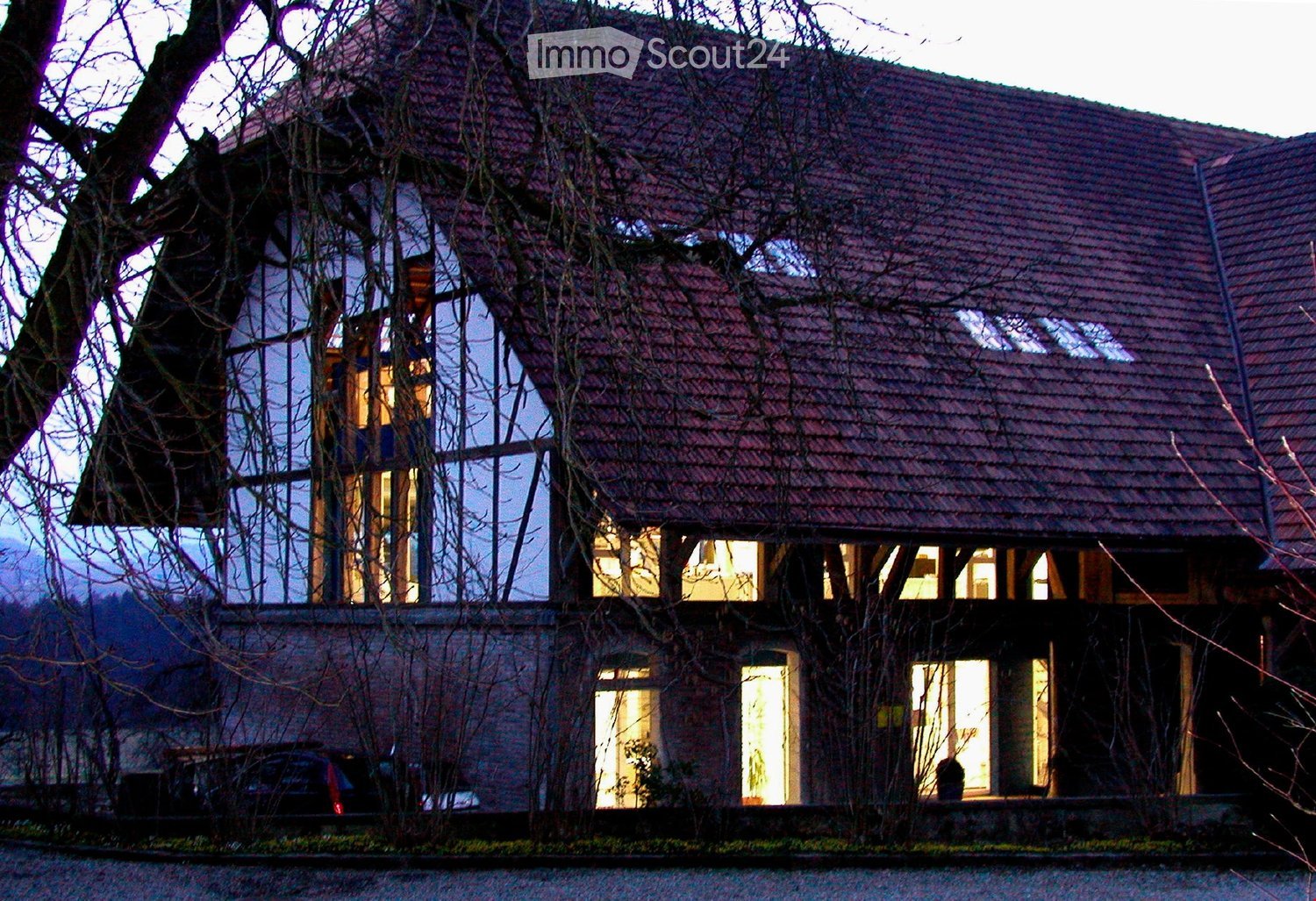 Two-story house with red-tiled roof, large windows, and visible interior lighting. The house features a brick lower level and a wooden upper level with a prominent tree integrated into the structure.