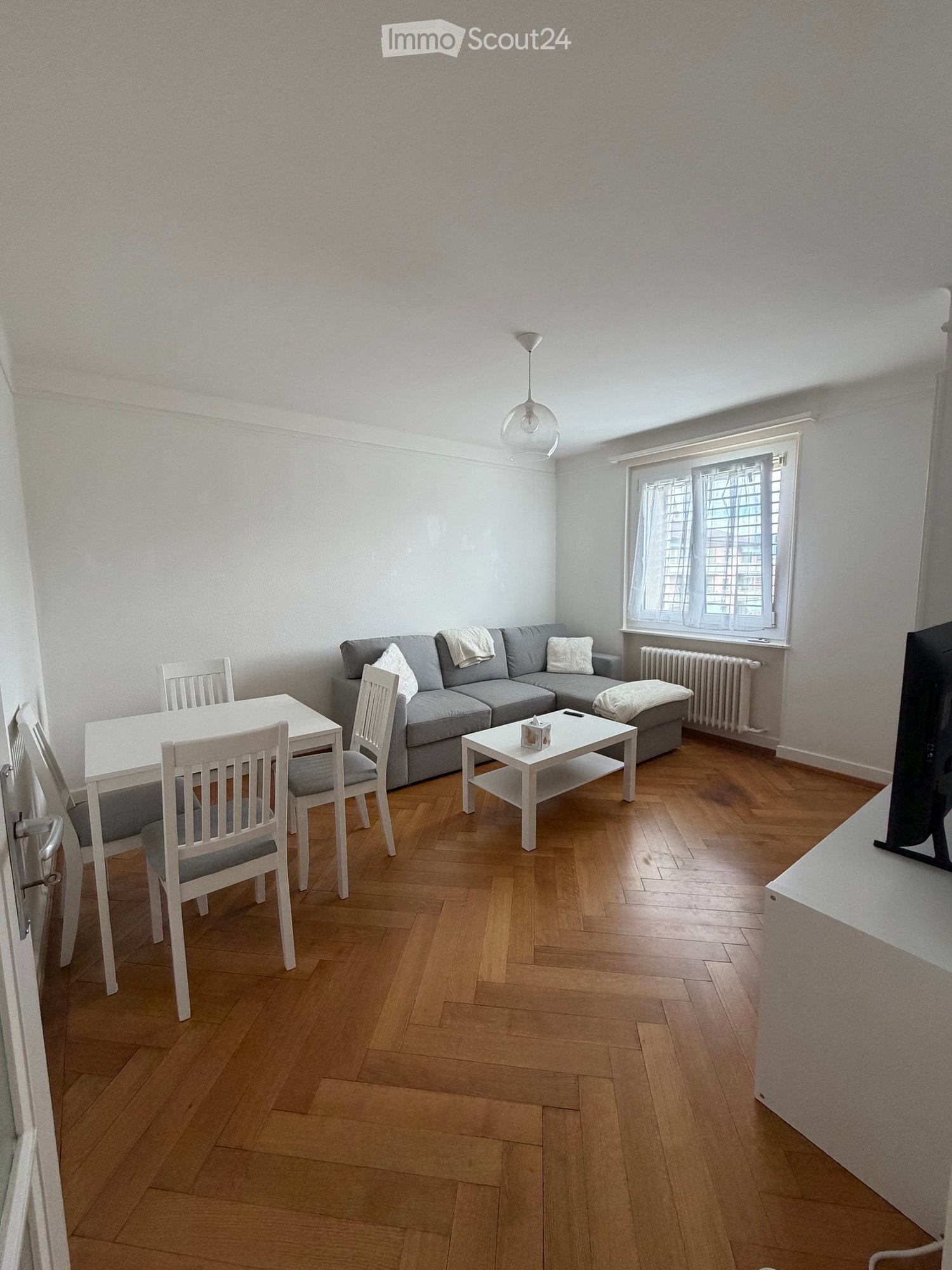 Living room with grey sectional sofa, white coffee table, white dining table with chairs, and a television on a stand.