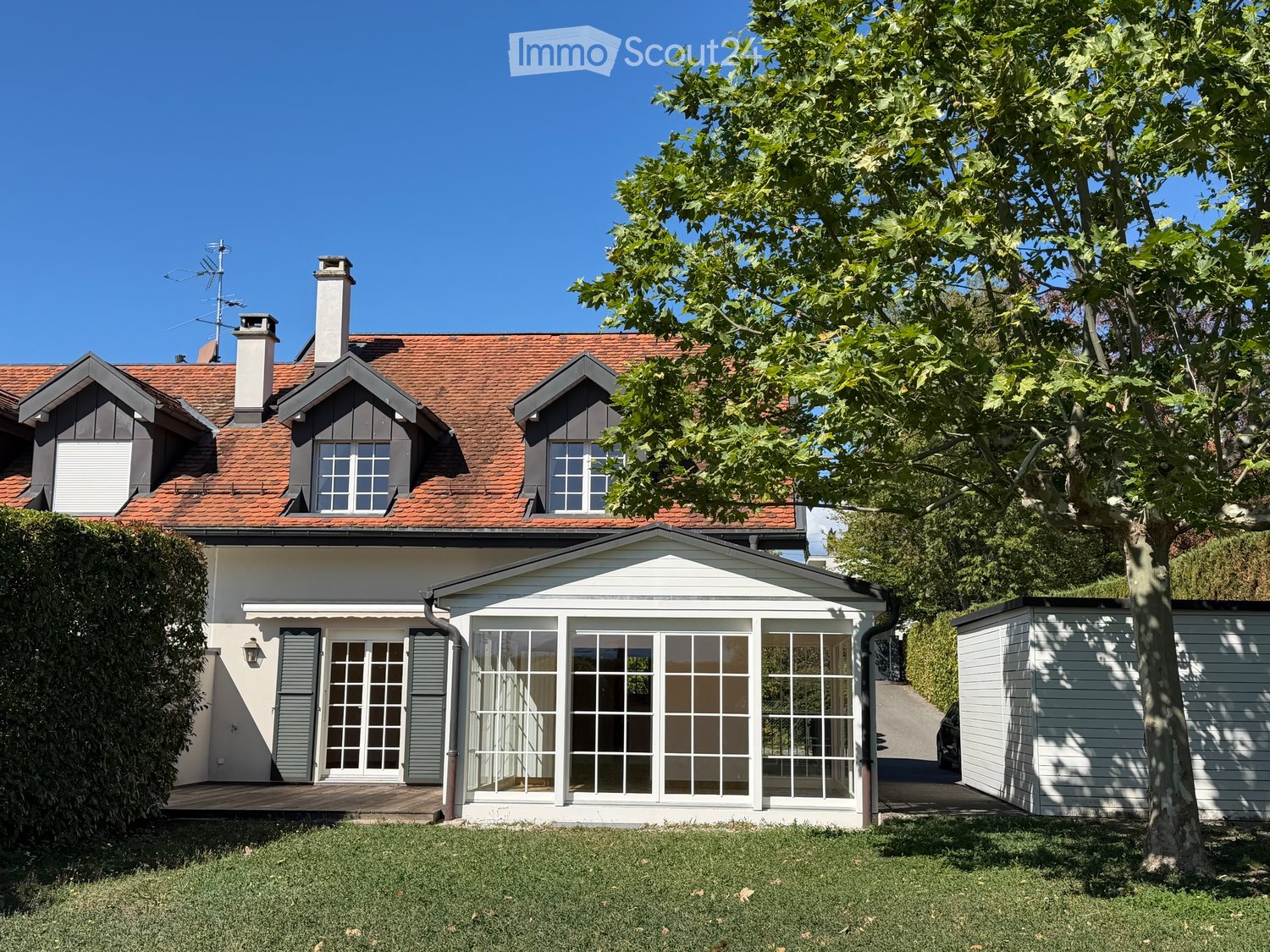 Two-story house with red-tiled roof, white walls, multiple chimneys, and large windows. The house features a well-maintained garden with a large tree and a small shed.