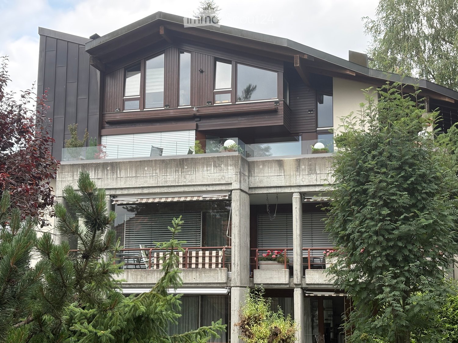 Modern two-story house with large windows, balconies, and surrounding greenery.