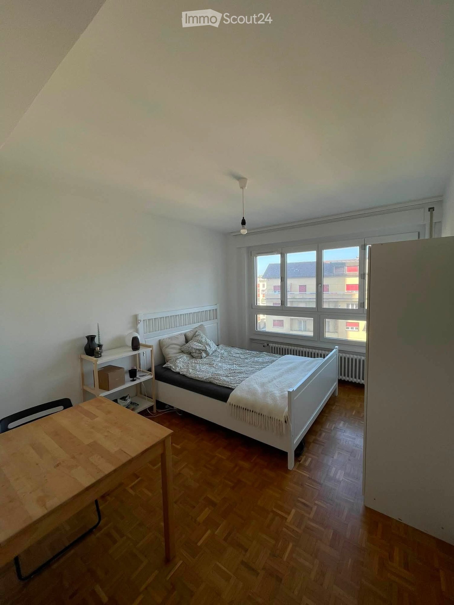 A cozy bedroom with a wooden bed, a small wooden table, a shelf with decorative items, and a window overlooking a cityscape.